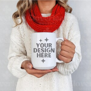 May include: White ceramic mug with a gray design that says "YOUR DESIGN HERE" with two small stars on either side of the text. The mug is being held by a person wearing a white sweater and a red scarf.