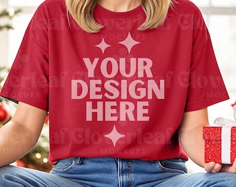 Könnte beinhalten: Ein rotes T-Shirt mit dem Text "YOUR DESIGN HERE" in Weiß. Das Shirt wird von einer Person getragen, die sitzt und eine rote Geschenkbox hält.