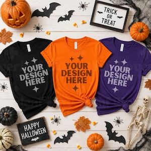 May include: Three short-sleeved crew neck t-shirts in black, orange, and purple, each featuring the text YOUR DESIGN HERE in a bold font. The shirts are arranged on a light wooden surface surrounded by pumpkins, bats, and Halloween decor.