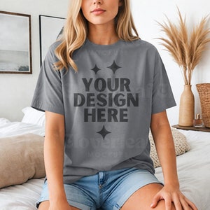 May include: A gray oversized t-shirt with short sleeves featuring the text "YOUR DESIGN HERE" in black. The shirt is worn by a person sitting on a bed.