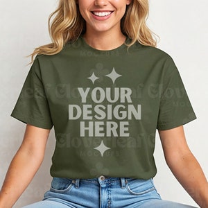 以下が含まれることがあります： 女性が、前面に「YOUR DESIGN HERE」という文字がプリントされた濃い緑色のTシャツを着ています。Tシャツは半袖で、クルーネックです。