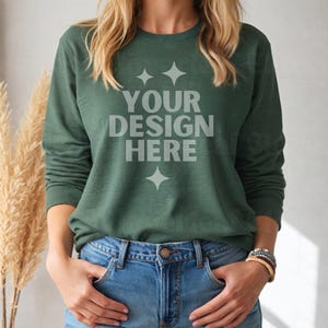 May include: A dark green long-sleeved shirt with the text "YOUR DESIGN HERE" printed on the front. The shirt is worn by a person with long blonde hair and is paired with blue jeans.