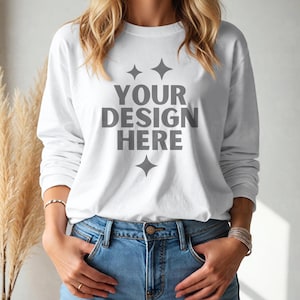 May include: A white long-sleeved t-shirt with the text "YOUR DESIGN HERE" printed on the front. The shirt is worn by a person with long blonde hair and blue jeans.