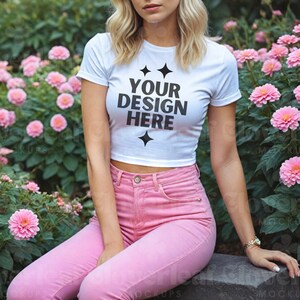 May include: A white crop top with short sleeves and the text "YOUR DESIGN HERE" printed on the front. The model is wearing pink jeans and is sitting in a garden setting.