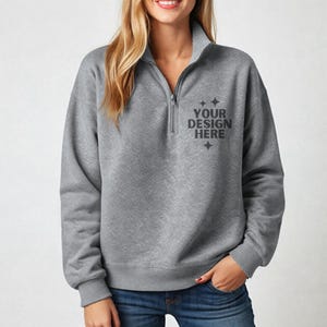 May include: A gray quarter-zip sweatshirt with the text "YOUR DESIGN HERE" printed on the chest. The sweatshirt has a high collar and long sleeves. The model is wearing blue jeans.