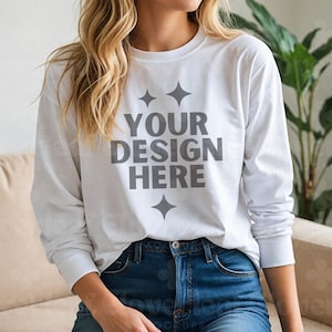 May include: A white long-sleeve t-shirt with the text "YOUR DESIGN HERE" in gray lettering. The shirt has a relaxed fit and is worn by a person with long blonde hair. The person is wearing blue jeans.