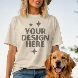 May include: Cream-colored t-shirt with the text "YOUR DESIGN HERE" in gray, with starburst graphics above and below the text. A golden retriever dog is in the foreground. The person is wearing blue jeans.