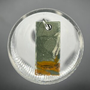 May include: A green, white, and yellow stone pendant with a silver bail. The stone has a rectangular shape and a smooth, polished surface.