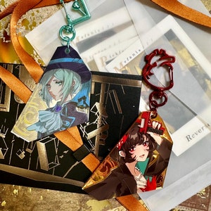 Puede incluir: Dos encantos de personajes con ilustraciones de estilo anime. El primer encanto es un personaje verde y azul con un sombrero y una gema azul en el pecho. El segundo encanto es un personaje de cabello castaño con una bandana roja y un arma roja y dorada. Ambos encantos están unidos a una cadena dorada con un cierre en forma de corazón.