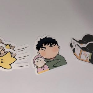 May include: A set of three cartoon stickers. One shows a bear hugging a star, another depicts a man holding a baby, and the last shows two figures reading a book.