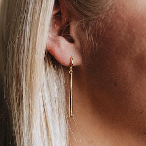 Puede incluir: Primer plano de la oreja de una persona con un pendiente dorado de aro. El pendiente es una barra fina y recta que cuelga de un aro pequeño.
