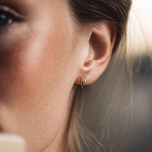 May include: A close-up shot of a person wearing a pair of small gold hoop earrings with a single, small diamond. The earrings are set against the person's skin, with a blurred background. The earrings are the focal point of the image.