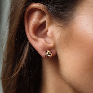 May include: A close-up of a gold-colored knot stud earring. The earring is small and round, with a textured surface. The earring is worn on an ear.