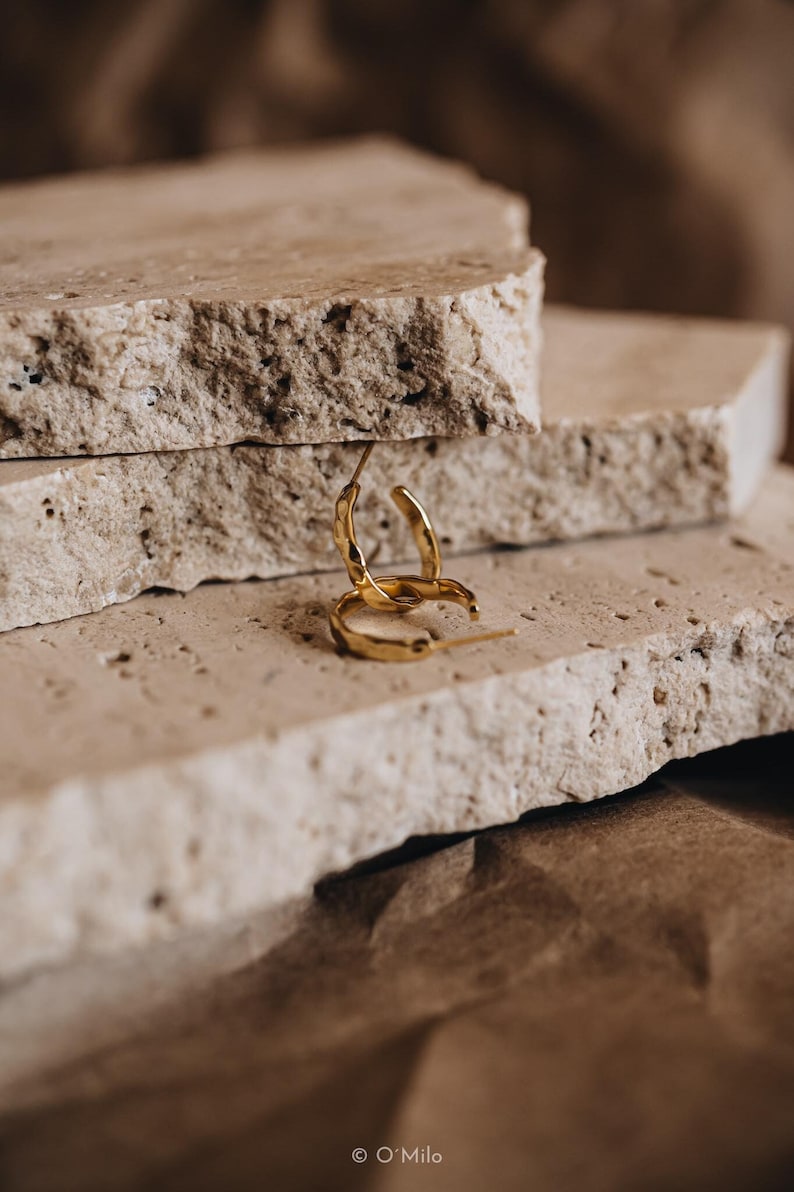 May include: Two gold hoop earrings with a textured surface, resting on a stack of light brown stone tiles.