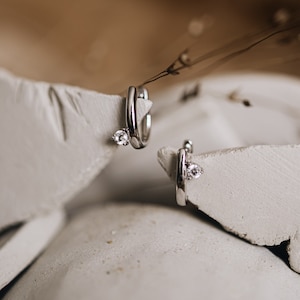 Puede incluir: Un par de pendientes de aro plateados, cada uno con una peque&ntilde;a gema transparente. Los pendientes se muestran sobre una superficie texturizada de color blanco roto. La imagen tiene una paleta de colores suave y neutra, con enfoque en joyer&iacute;a.