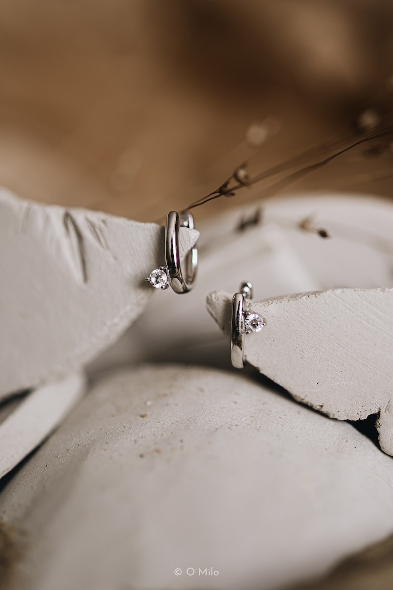 Puede incluir: Un par de pendientes de aro plateados, cada uno con una peque&ntilde;a gema transparente. Los pendientes se muestran sobre una superficie texturizada de color blanco roto. La imagen tiene una paleta de colores suave y neutra, con enfoque en joyer&iacute;a.