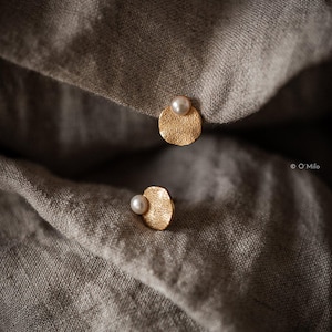 May include: Gold-coloured stud earrings with a textured finish, each featuring a small, round, off-white pearl. The earrings are presented on a grey, textured fabric.