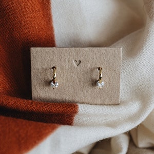 Può includere: Un paio di orecchini a cerchio dorati con un piccolo ciottolo trasparente su ogni cerchio. Gli orecchini sono esposti su una carta marrone con un cuore ritagliato.