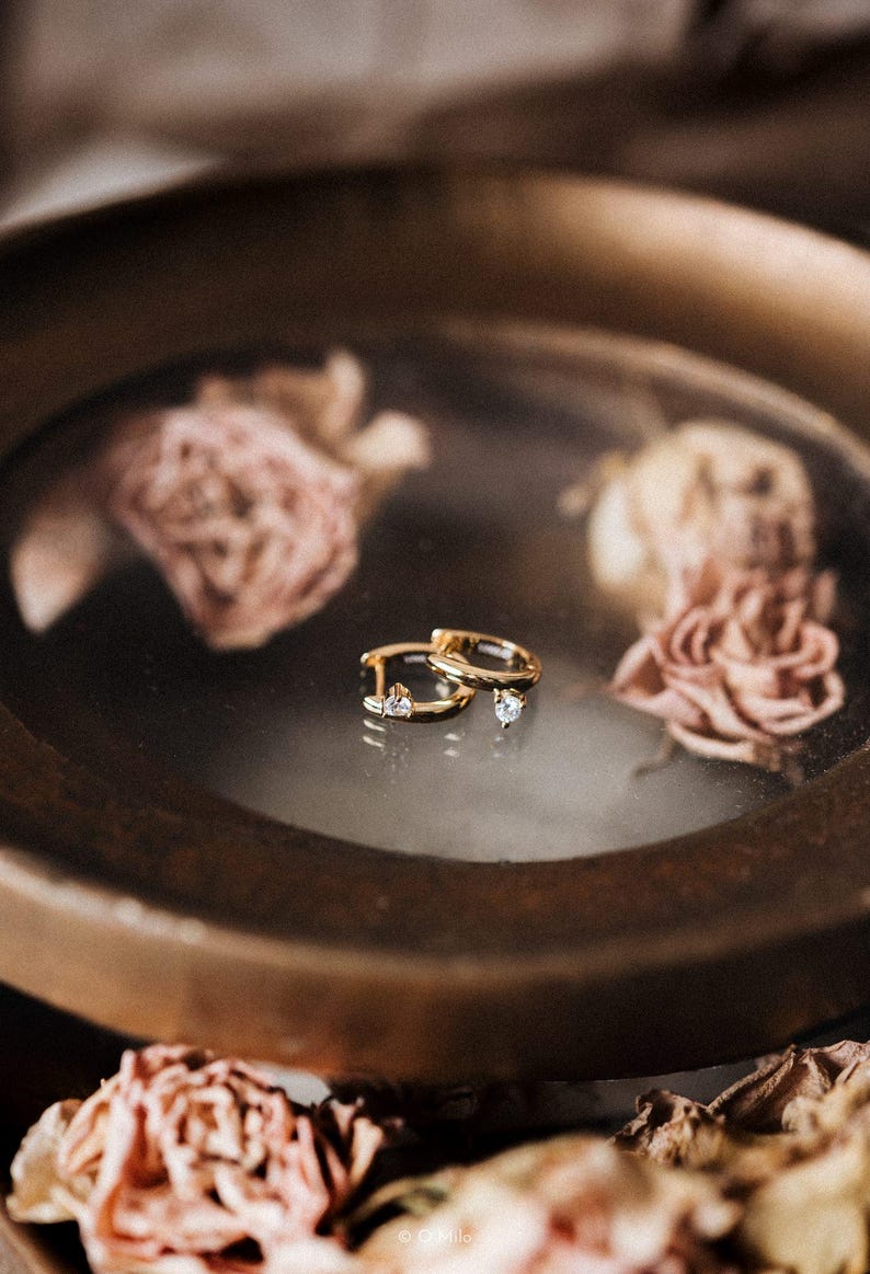 Puede incluir: Un par de pendientes de aro dorados con peque&ntilde;as piedras preciosas transparentes. Los pendientes se exhiben sobre una superficie reflectante dentro de un plato de color bronce, rodeados de rosas rosas secas. Los pendientes tienen un dise&ntilde;o cl&aacute;sico y elegante.