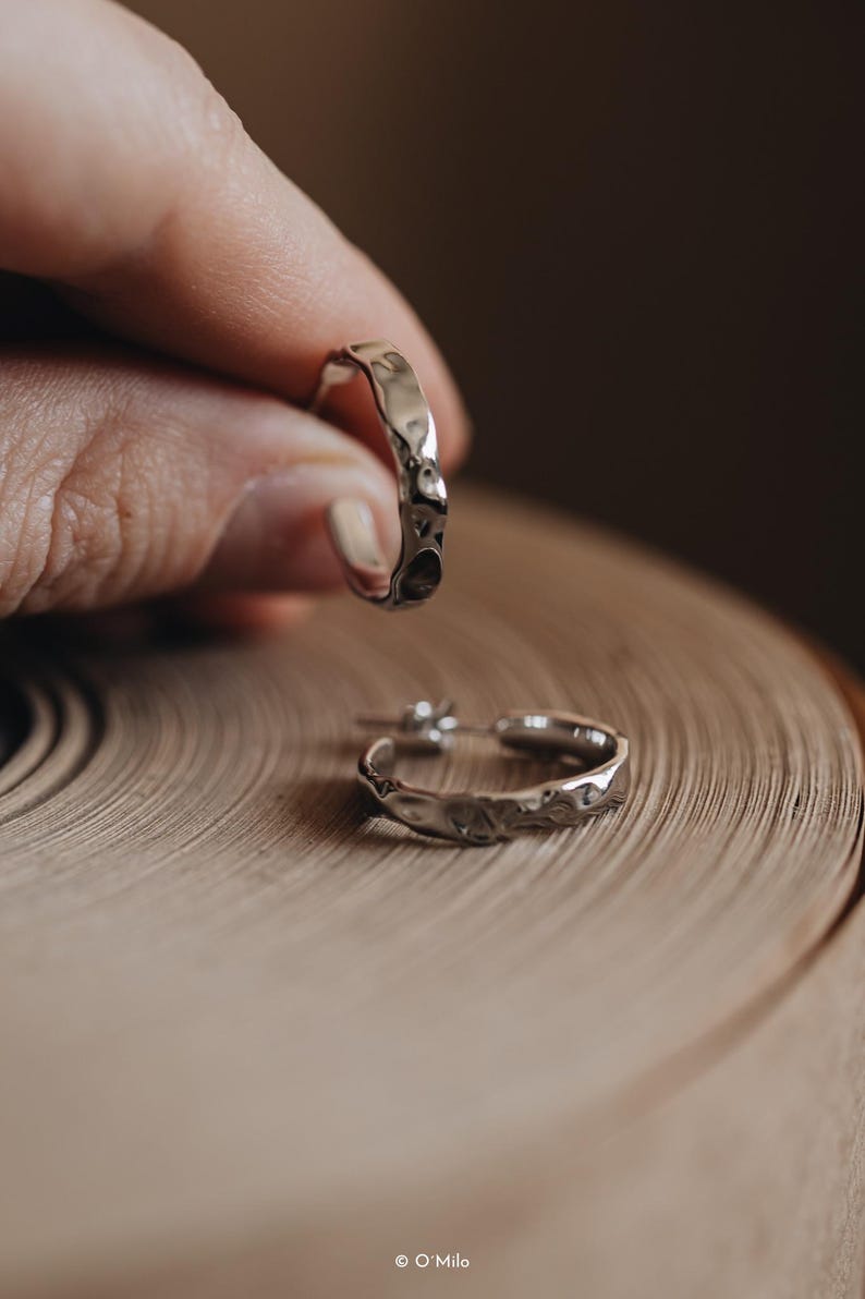 May include: A pair of silver hoop earrings with a textured, hammered finish. The earrings are shown on a wooden surface.