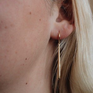Puede incluir: Un par de pendientes largos y finos de oro con una punta puntiaguda. Los pendientes cuelgan del lóbulo de la oreja de una persona con cabello rubio.