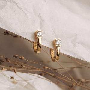 Könnte beinhalten: Ein Paar goldfarbene Creolen, die jeweils mit einem einzelnen runden, klaren Stein an der Oberseite besetzt sind. Das Innenband jedes Ohrrings ist deutlich mit der Aufschrift 925 versehen, was auf Sterlingsilber mit Goldbeschichtung hinweist.