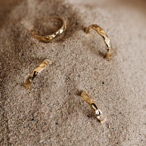 May include: Four gold hammered rings on a sandy surface. The rings are all different sizes and have a textured finish.