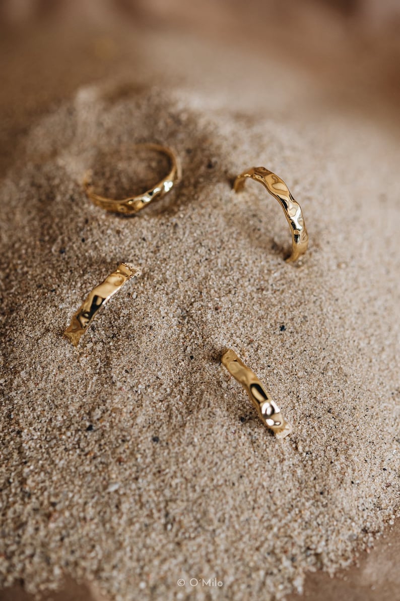 May include: Four gold hammered rings on a sandy surface. The rings are all different sizes and have a textured finish.
