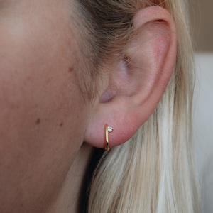 Puede incluir: Primer plano de un pendiente de aro dorado con una peque&ntilde;a gema transparente. El pendiente se lleva en la oreja de una persona, con pelo rubio visible en el fondo. Dise&ntilde;o minimalista.