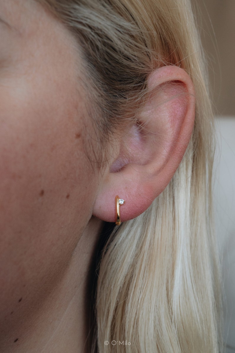 Puede incluir: Primer plano de un pendiente de aro dorado con una peque&ntilde;a gema transparente. El pendiente se lleva en la oreja de una persona, con pelo rubio visible en el fondo. Dise&ntilde;o minimalista.