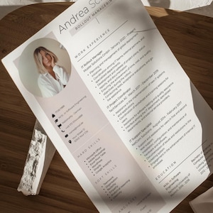 Op de afbeelding: Een cv getiteld "Andrea Schiller, Rollout Manager" met een foto, werkervaring, vaardigheden en opleidingsdetails. Het cv staat op wit papier met een roze accent, geplaatst op een houten ondergrond met een wit decoratief stuk.