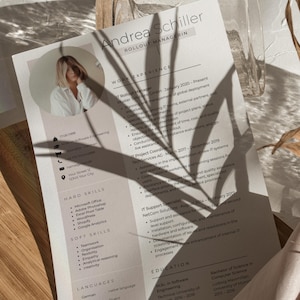 May include: A resume with the name "Andrea Schiller" and the title "Rollout Managerin" is displayed on a wooden surface. The resume includes sections for work experience, hard skills, soft skills, and languages. The resume is partially covered by shadows.