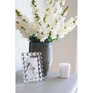 May include: A clear glass picture frame with a beaded border sits on a gray surface next to a white candle and a vase of white flowers.