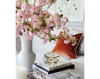 May include: A white ceramic vase filled with pink cherry blossom branches sits on a coffee table with a stack of books and a clear crystal decorative object. A burnt orange velvet pillow with a white border sits on a white couch.