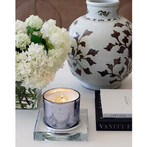 May include: A silver candle with a blue and white design sits on a glass candle holder. The candle is lit and there are three wicks. The candle is on a white surface with a white vase of white flowers and a black book with white text.