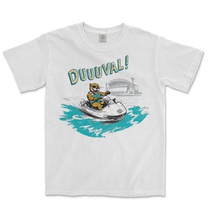 May include: White t-shirt featuring a cartoon illustration of a jaguar riding a jet ski with the text "DUUUVAL!" printed on the front.