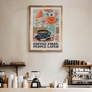 May include: A framed art print with a gold frame, featuring a coffee cup and floral arrangement. The print has the text "COFFEE FIRST, PEOPLE LATER". The image is displayed in a coffee shop setting, with coffee-making equipment visible.