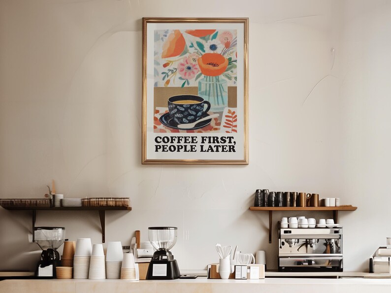May include: A framed art print with a gold frame, featuring a coffee cup and floral arrangement. The print has the text "COFFEE FIRST, PEOPLE LATER". The image is displayed in a coffee shop setting, with coffee-making equipment visible.