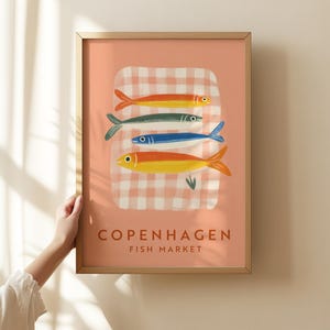 Könnte beinhalten: Gerahmter Kunstdruck mit vier farbenfrohen Fischillustrationen auf rosa Hintergrund mit Karomuster. Der Druck enthält den Text "COPENHAGEN FISH MARKET". Der Rahmen hat eine hellbraune Farbe.