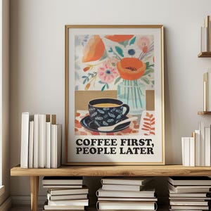 May include: A framed art print with a gold border featuring a floral and coffee-themed design. The artwork includes a cup of coffee, flowers, and the text "COFFEE FIRST, PEOPLE LATER". The print is displayed on a wooden shelf with books.