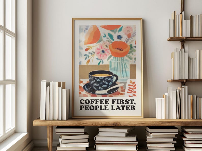 May include: A framed art print with a gold border featuring a floral and coffee-themed design. The artwork includes a cup of coffee, flowers, and the text "COFFEE FIRST, PEOPLE LATER". The print is displayed on a wooden shelf with books.