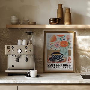May include: A kitchen scene featuring a stainless steel espresso machine, a framed art print with a floral and coffee cup design, and a white coffee cup on a saucer. The art print reads "COFFEE FIRST, PEOPLE LATER."