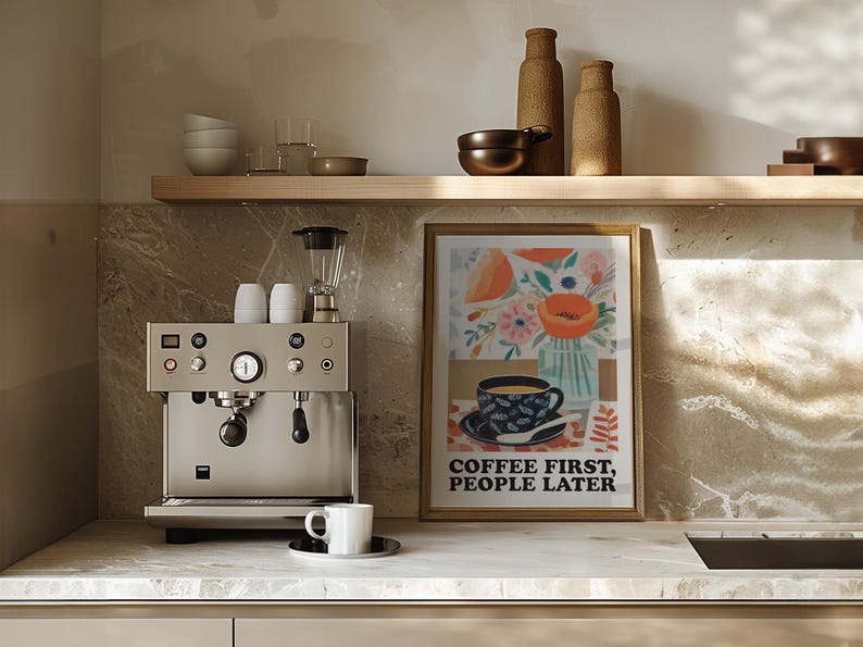 May include: A kitchen scene featuring a stainless steel espresso machine, a framed art print with a floral and coffee cup design, and a white coffee cup on a saucer. The art print reads "COFFEE FIRST, PEOPLE LATER."