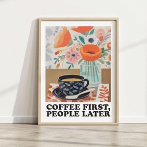 May include: A framed art print featuring a coffee cup and flowers. The cup and saucer are navy blue with a floral pattern. The print includes the text "COFFEE FIRST, PEOPLE LATER". The background includes a vase of flowers and a floral pattern.