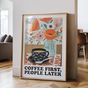 May include: Framed art print featuring a coffee cup with a floral design, a vase of flowers, and the text "COFFEE FIRST, PEOPLE LATER." The artwork has a gold frame and is leaning against a white wall in a room with hardwood floors.