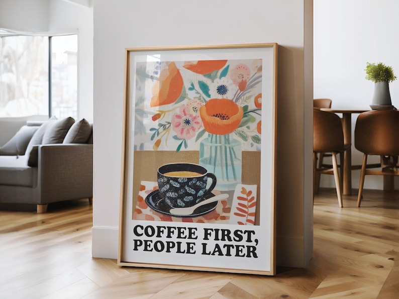 May include: Framed art print featuring a coffee cup with a floral design, a vase of flowers, and the text "COFFEE FIRST, PEOPLE LATER." The artwork has a gold frame and is leaning against a white wall in a room with hardwood floors.