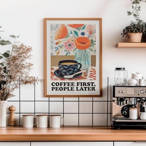May include: A framed art print with the text "COFFEE FIRST, PEOPLE LATER" hangs above a wooden countertop. The print features a coffee cup, flowers, and a vase. A coffee machine and mugs are on the counter.