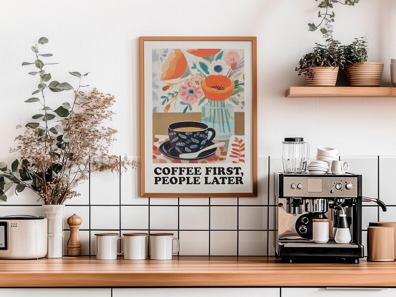 May include: A framed art print with the text "COFFEE FIRST, PEOPLE LATER" hangs above a wooden countertop. The print features a coffee cup, flowers, and a vase. A coffee machine and mugs are on the counter.