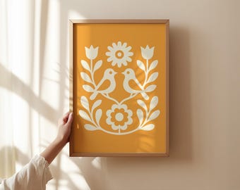 May include: Framed art print featuring a symmetrical design of two birds, flowers, and foliage in a cream color against a mustard yellow background. The artwork is in a wooden frame and is displayed on a neutral wall.