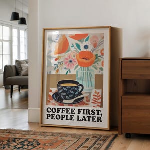 May include: Framed art print featuring a coffee cup with a floral design, a vase of flowers, and the text "COFFEE FIRST, PEOPLE LATER." The artwork has a warm color palette with orange, blue, and brown tones. The print is displayed in a room with a rug and wooden furniture.